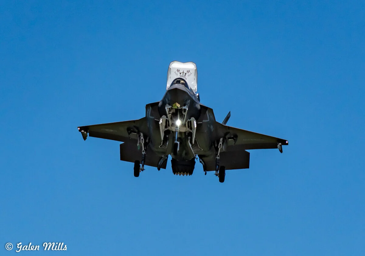 Fighter jet in flight against a clear blue sky, viewed from directly below, showing its undercarriage and landing gear.