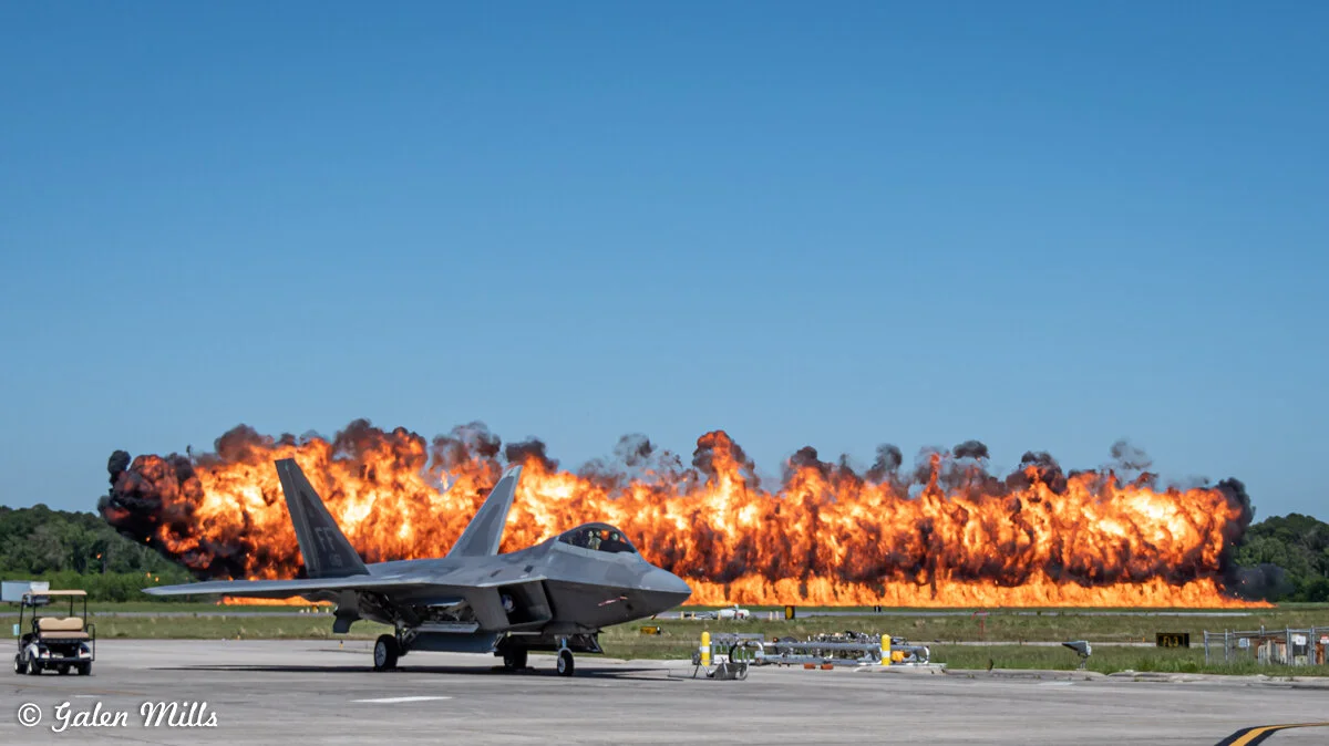 A fighter jet on a runway with a large explosion in the background.
