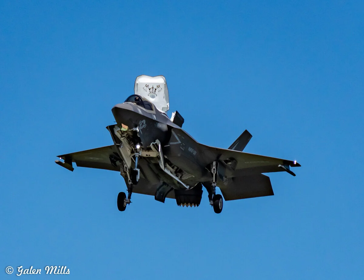 F-35 fighter jet flying with landing gear down against a clear blue sky.
