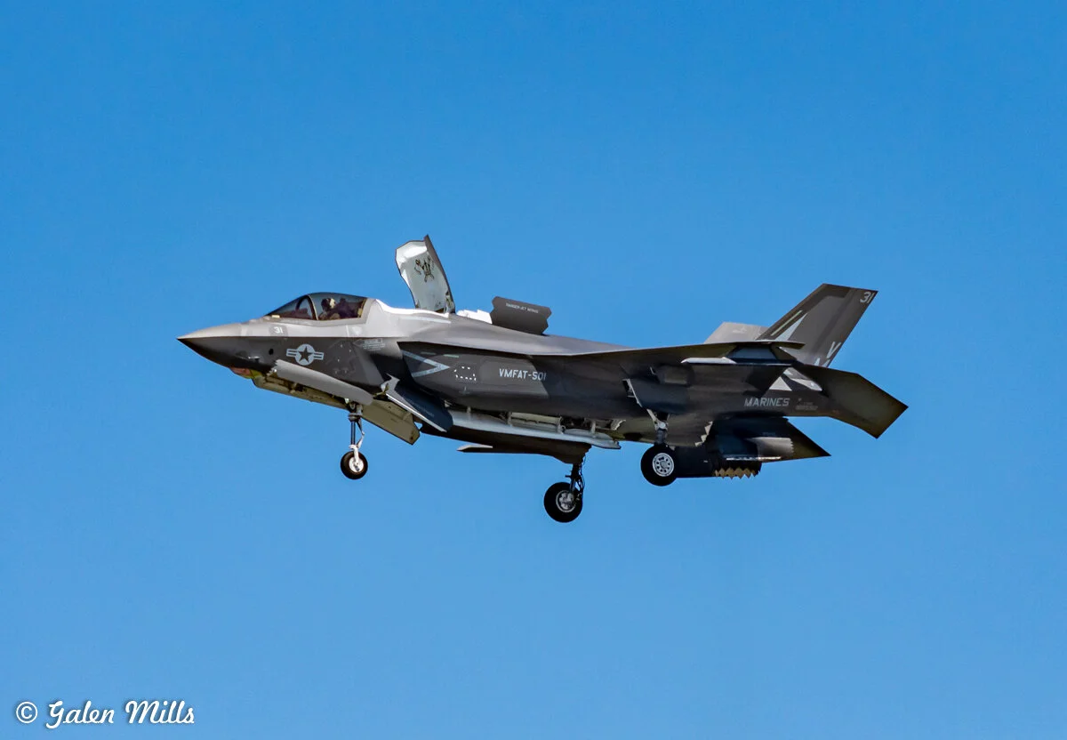 Military jet fighter in flight with landing gear deployed against clear blue sky.