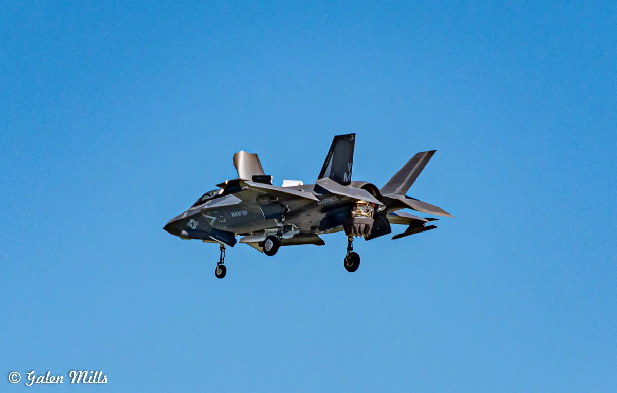 F-35B fighter jet flying with landing gear down against clear blue sky.