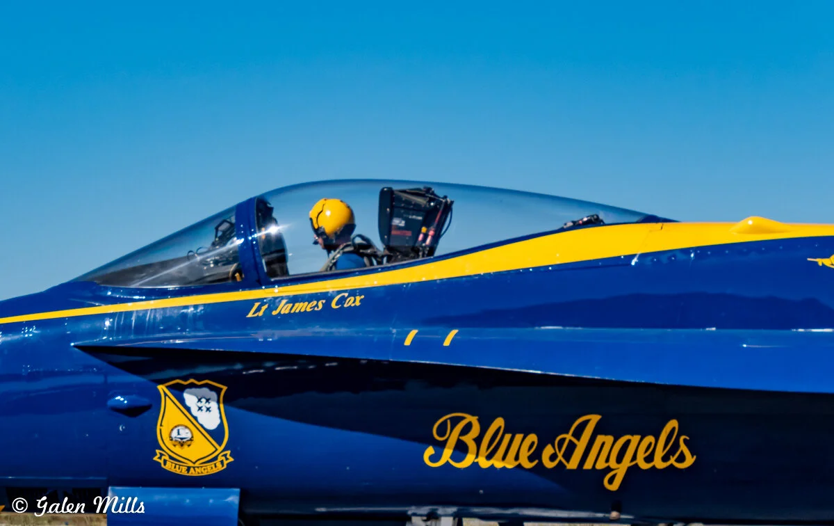A Blue Angels jet with a pilot, featuring a yellow helmet, inside the cockpit. The jet has "Lt James Cox" and "Blue Angels" inscribed on the side. The sky is clear blue in the background.