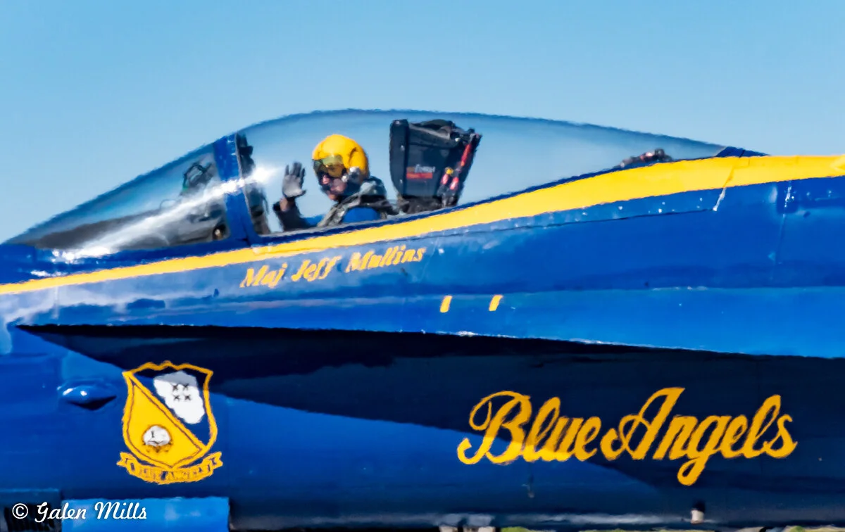 Blue Angels jet with pilot waving, cockpit with visible helmet, aircraft labeled 'Maj Jeff Mullins' and 'Blue Angels', blue and yellow color scheme.