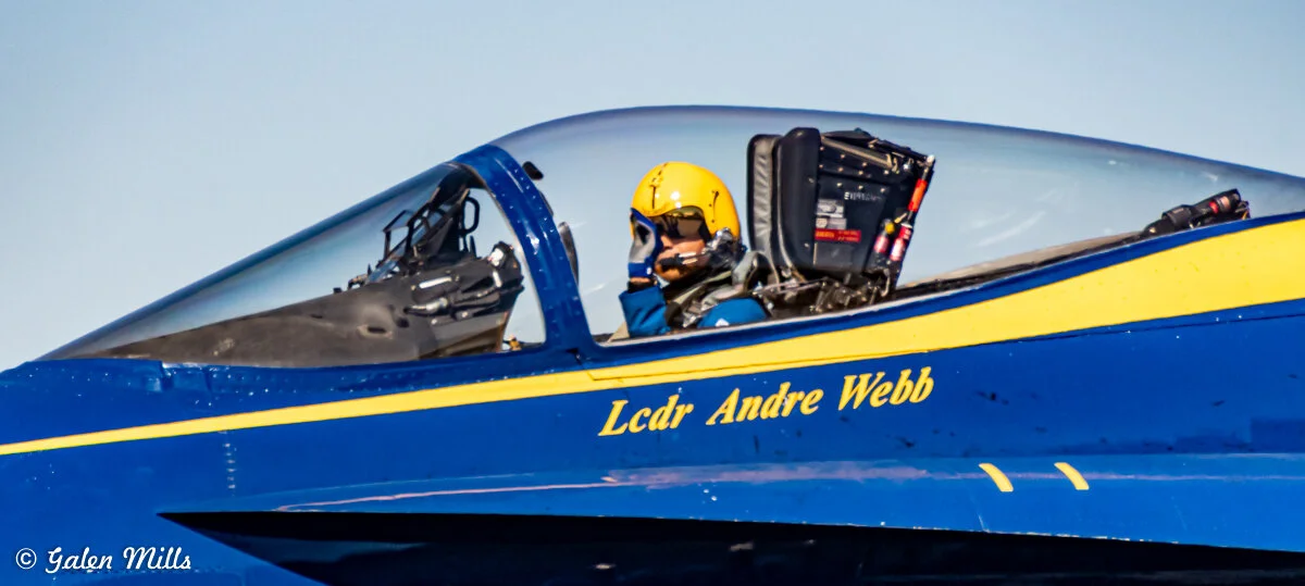 Close-up view of a Blue Angels pilot in a cockpit, wearing a yellow helmet and oxygen mask. The name 'Lcdr Andre Webb' is painted on the aircraft. Photo by Galen Mills.