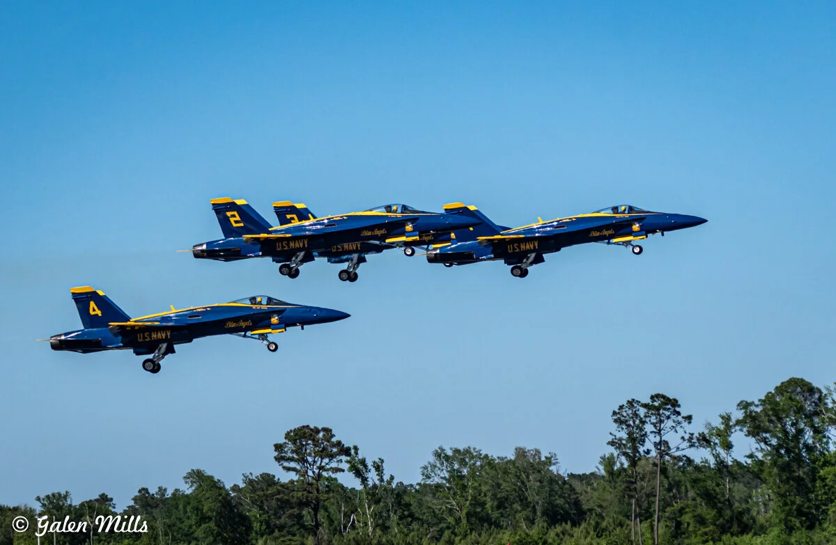 Four U.S. Navy Blue Angels jets flying in formation against a clear blue sky, with trees visible below.