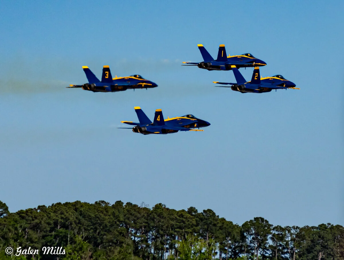 Four military jets in a formation flying over a forested area against a clear blue sky.