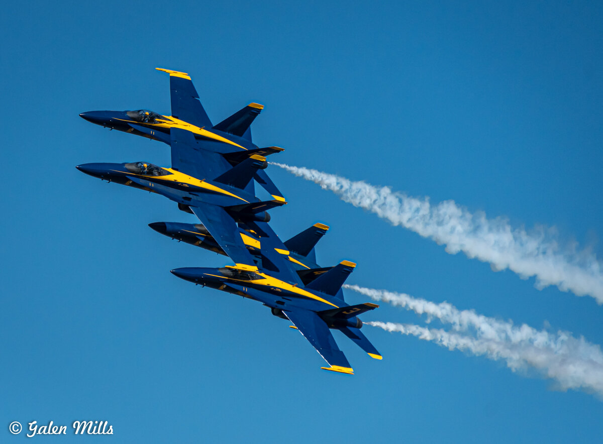Four fighter jets in close formation with blue and yellow markings, performing an aerial maneuver against a clear blue sky.