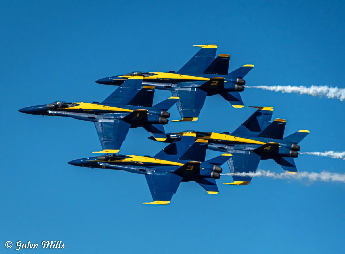 A group of four blue and yellow fighter jets performing an aerial formation against a clear blue sky.