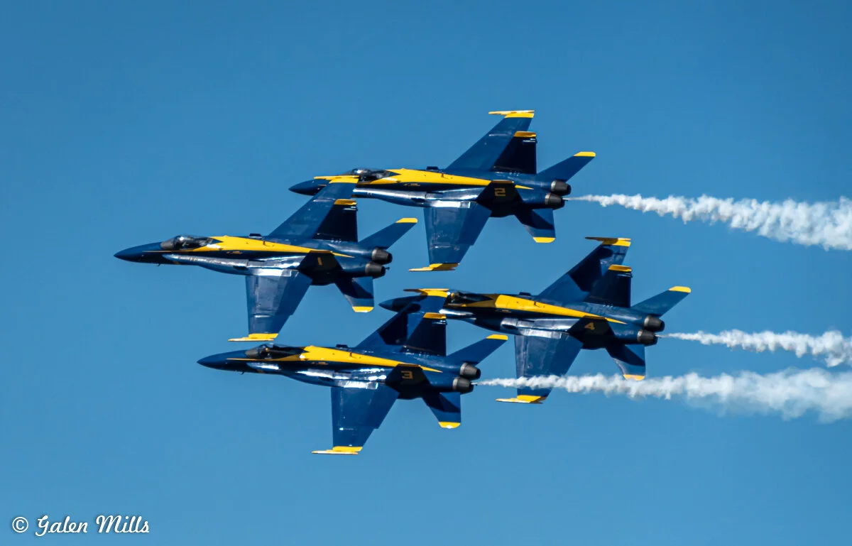 Four Blue Angels jets flying in a tight formation with smoke trails against a clear blue sky.