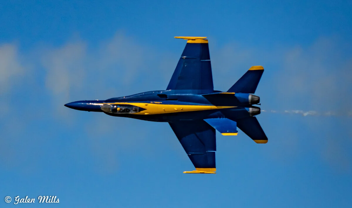 A Blue Angels jet performing an aerial maneuver against a clear blue sky.