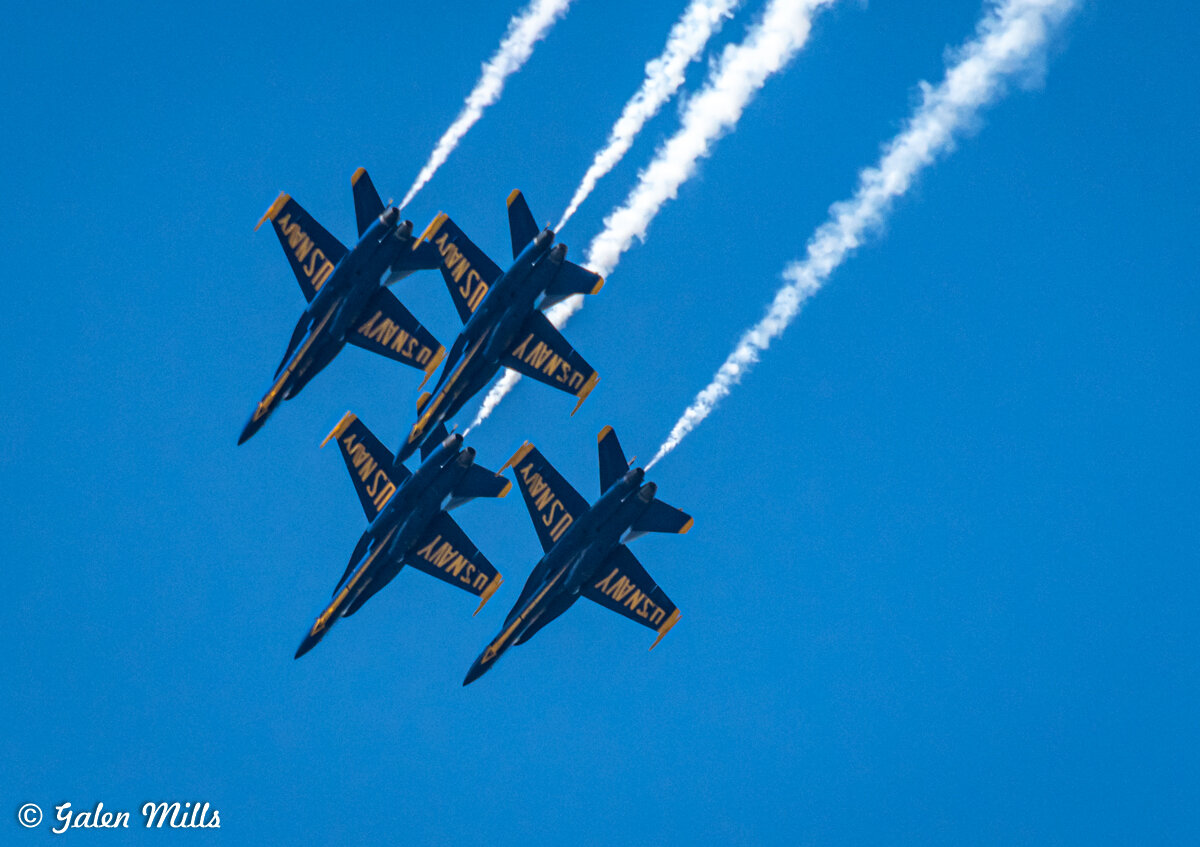 Four US Navy Blue Angels jets flying in formation with contrails against a clear blue sky.