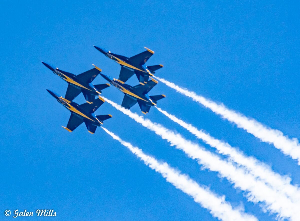 Four fighter jets flying in a diamond formation with white contrails against a blue sky.