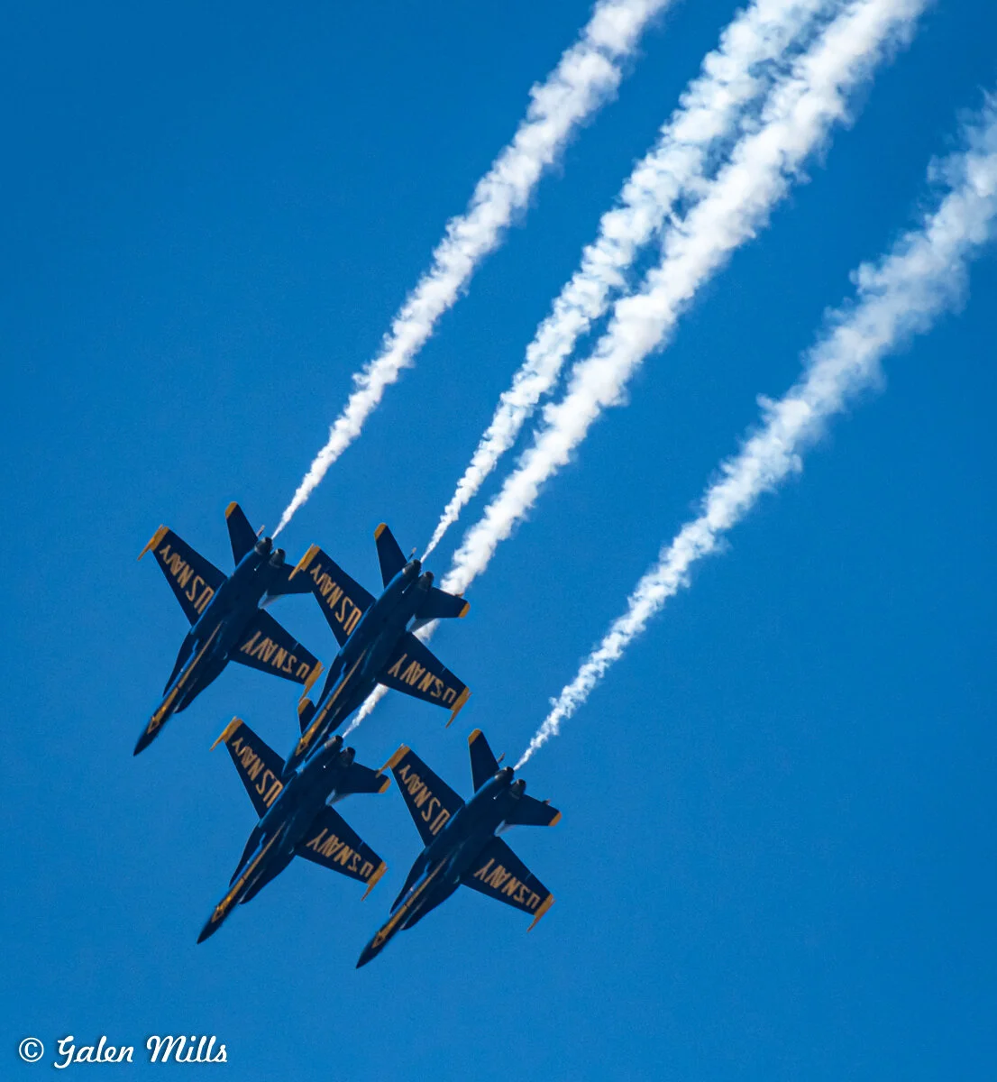 Blue Angels performing aerial maneuver with smoke trails against clear blue sky