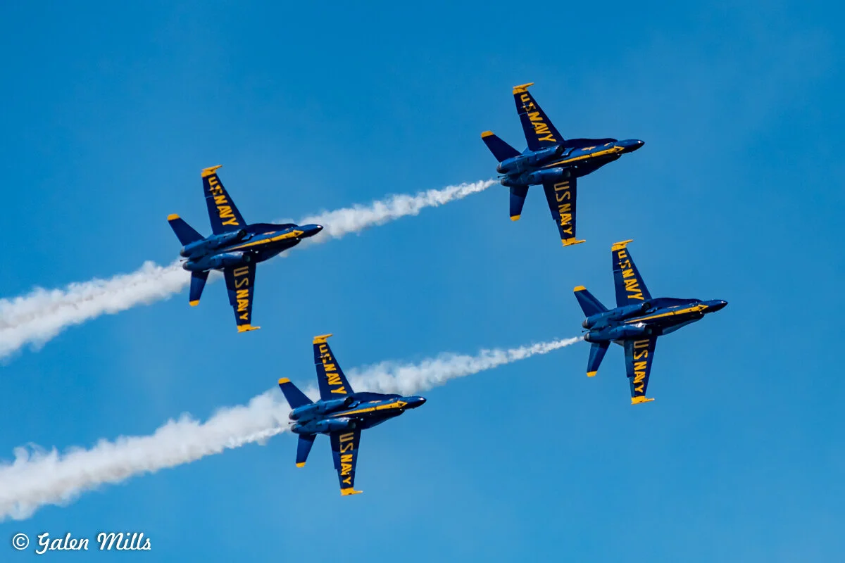 Four U.S. Navy Blue Angels jets performing an aerial maneuver with white smoke trails against a clear blue sky.