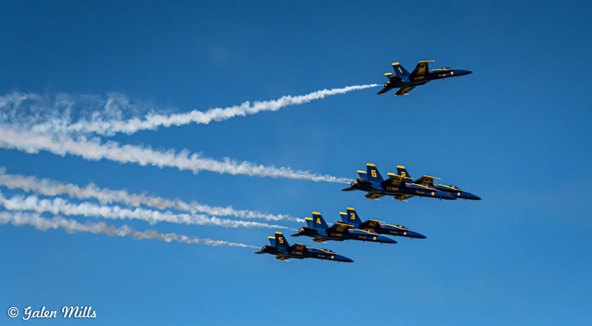 A formation of six U.S. Navy Blue Angels jets flying in a clear blue sky with smoke trails.