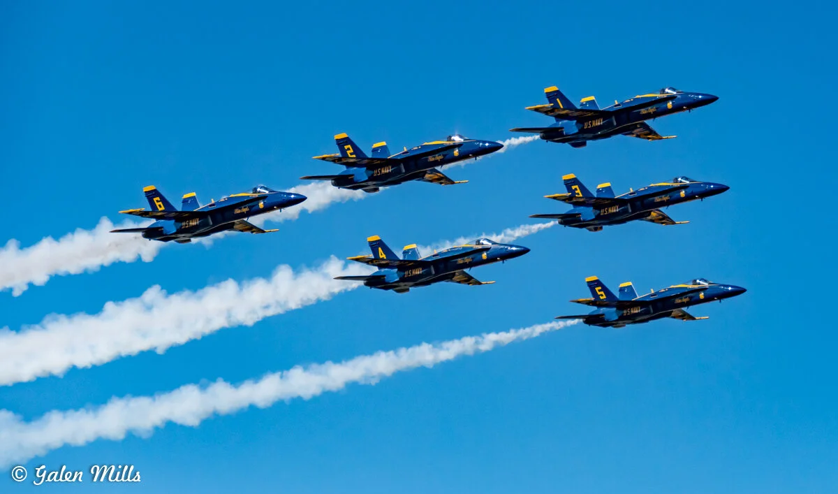 Six U.S. Navy Blue Angels jets flying in formation against a clear blue sky with smoke trails.