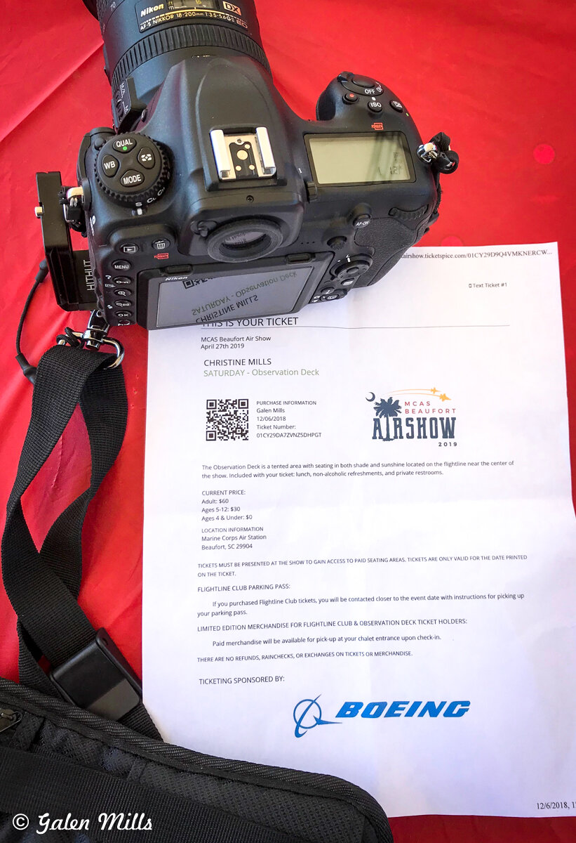 A Nikon camera with a zoom lens rests on a red surface next to an airshow ticket. The ticket includes details for the MCAS Beaufort Air Show on April 27, 2019, with observation deck seating. It shows purchase information and pricing, sponsored by Boe