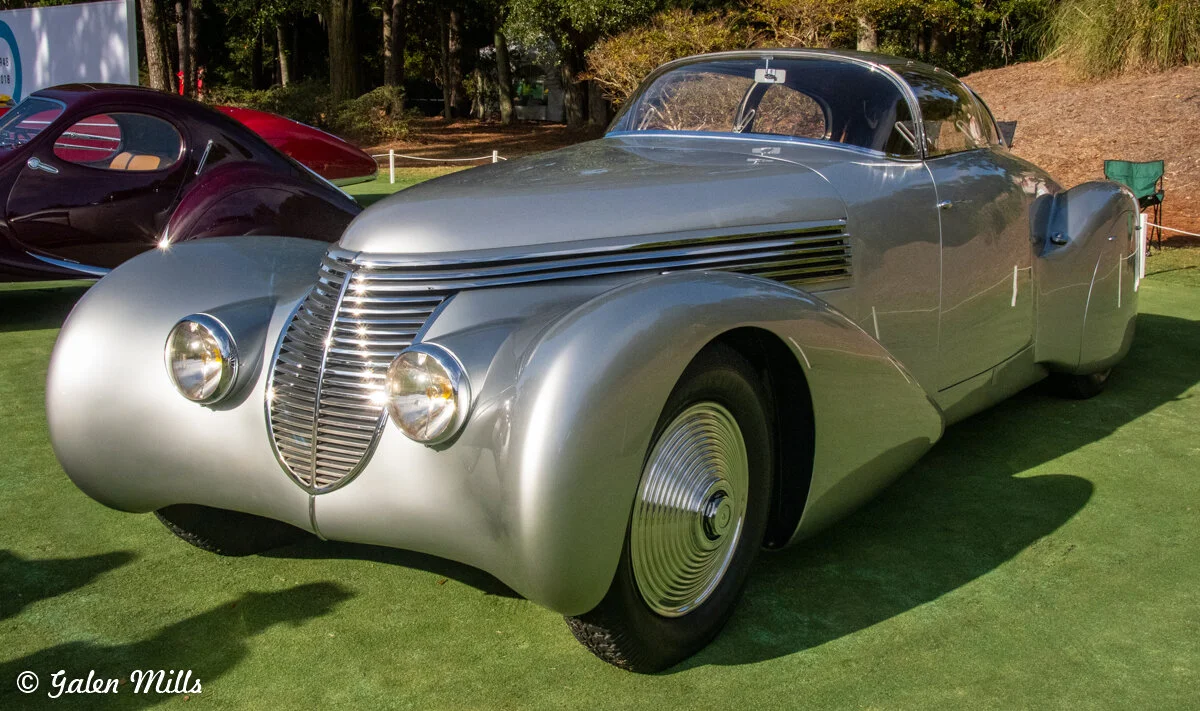 Vintage silver streamlined car with art deco design at a car show, featuring round headlamps and a distinctive grill.