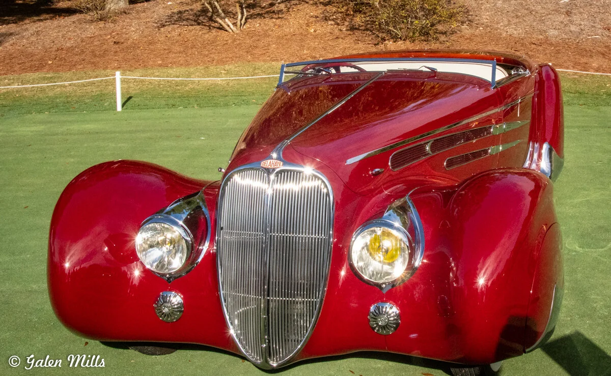 Red vintage car at a car show with a shiny chrome grille and distinctive fender design.
