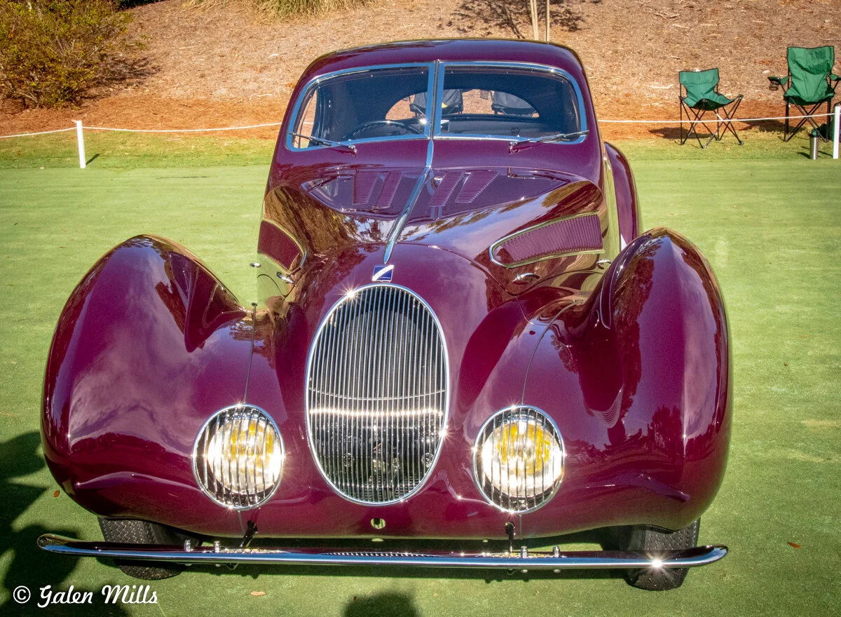 Vintage maroon car with rounded fenders and chrome grille at a car show, parked on grass.