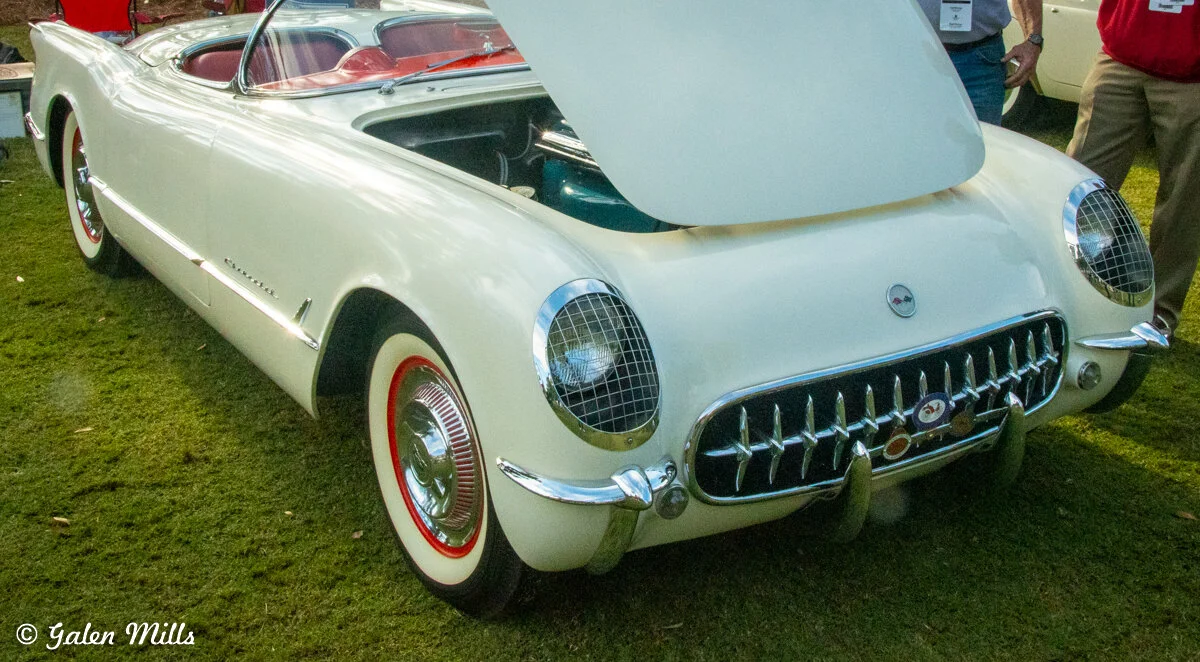 Vintage white convertible car with an open hood, featuring a classic chrome grille and red interior, parked on grass with people nearby.