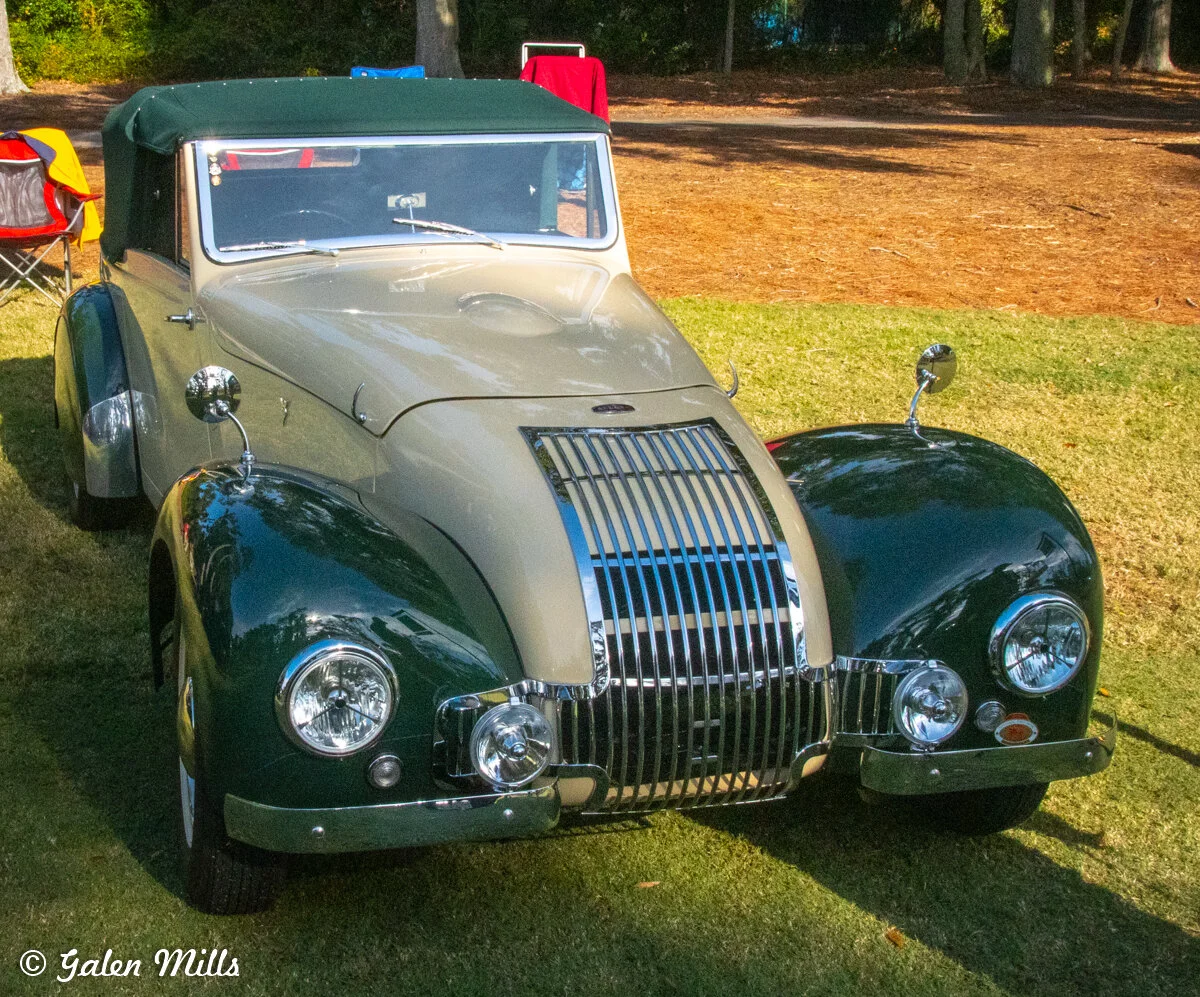 Classic vintage car with green and beige exterior on grassy area, surrounded by folding chairs.