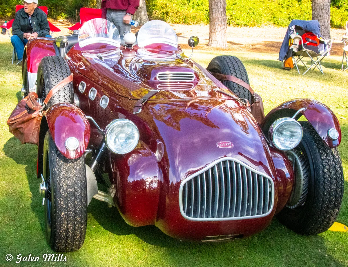Vintage maroon Allard sports car with exposed wheels and leather straps, parked on grass with people sitting nearby and trees in the background.