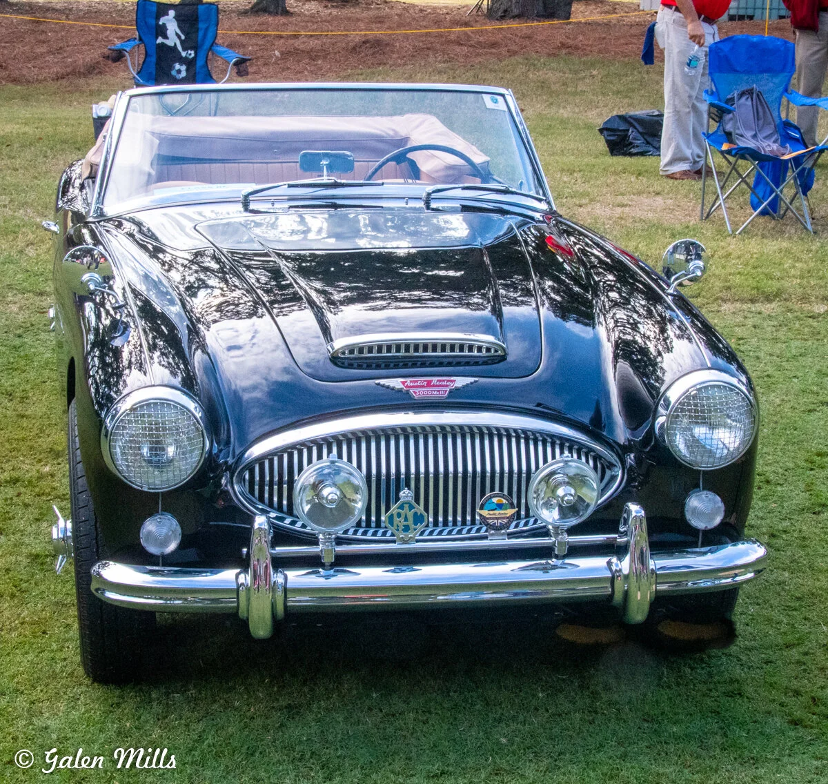 Classic black convertible car front view with chrome details, on grassy area, with chairs and people in the background.