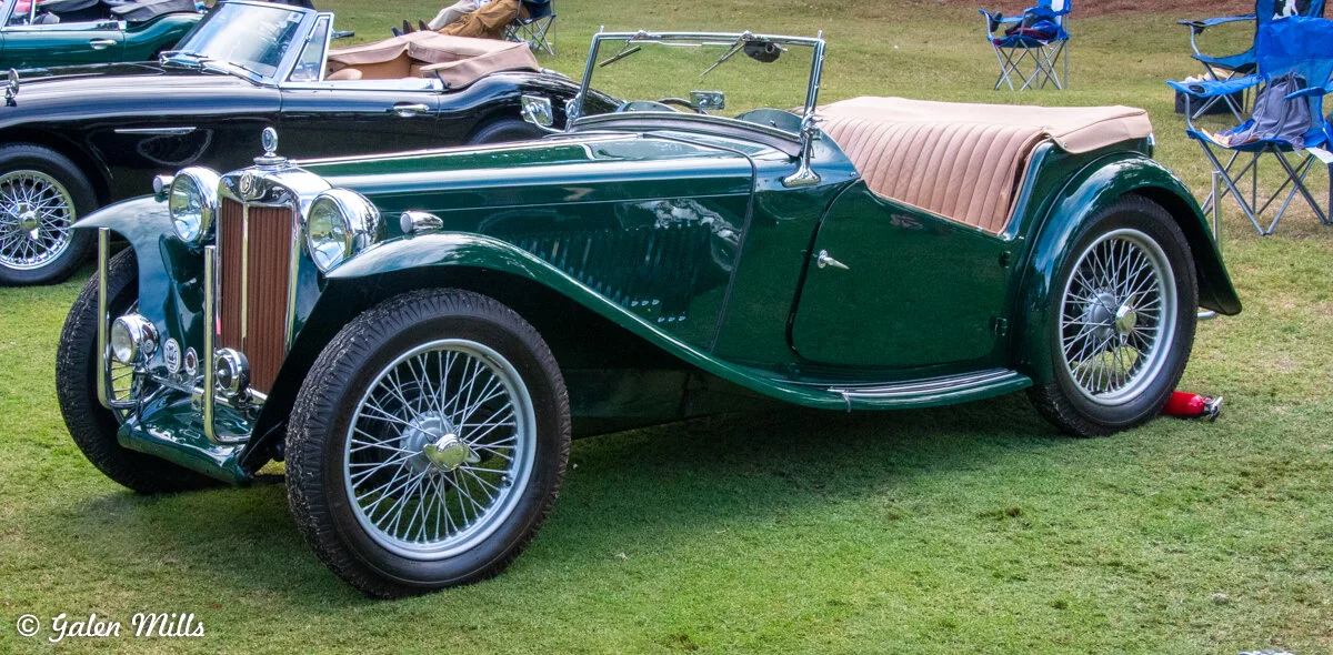 Vintage green convertible car on display in a grassy area with other classic cars and folding chairs nearby.