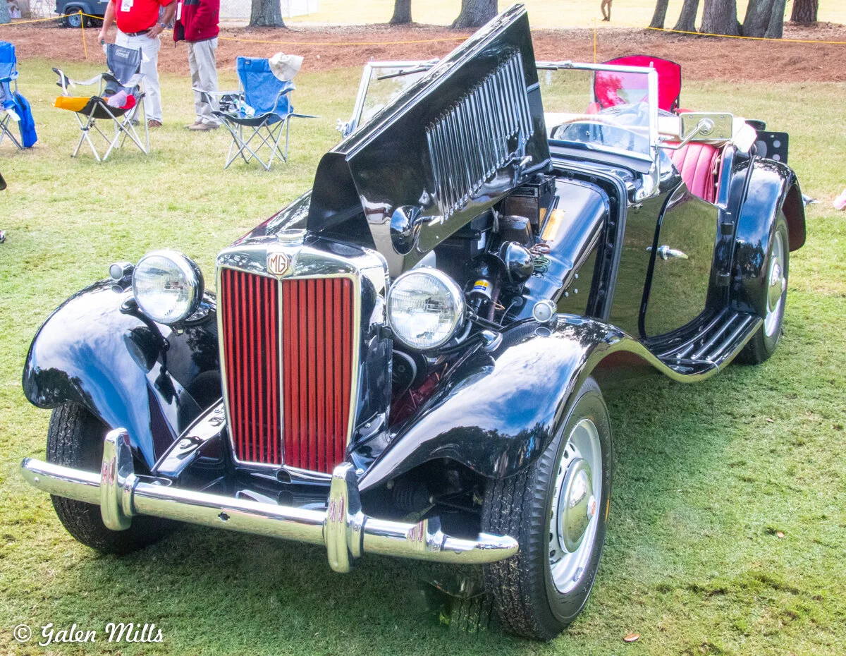 Classic black MG convertible with open hood on grass, surrounded by people and folding chairs.