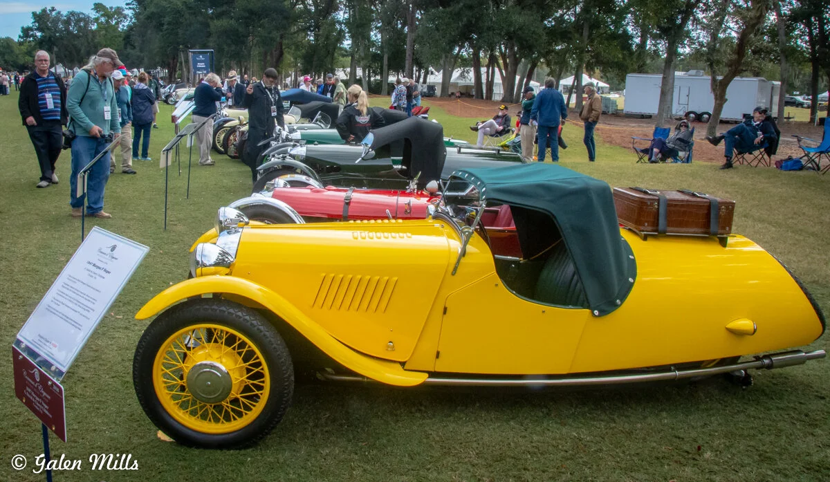Vintage yellow sports car displayed at a car show, surrounded by other classic cars and people admiring them in a park setting.