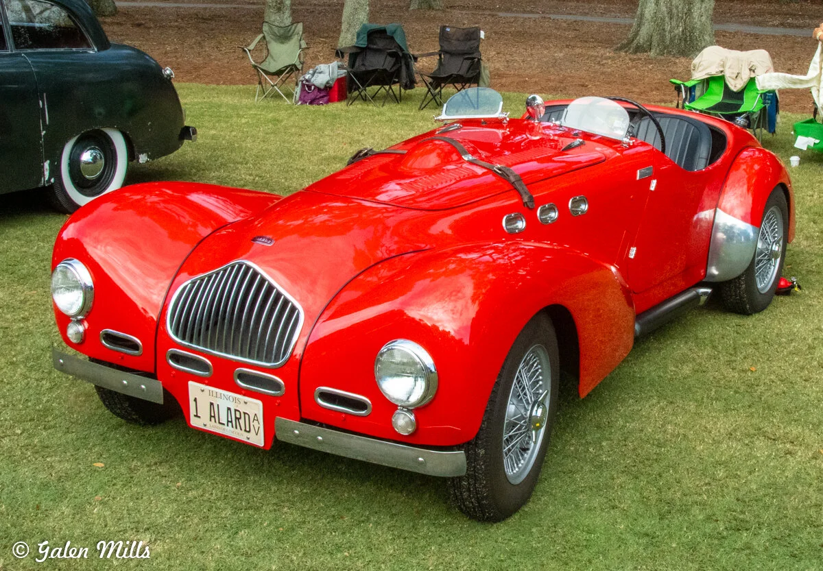 Red vintage sports car parked on grass with other cars and chairs in the background.