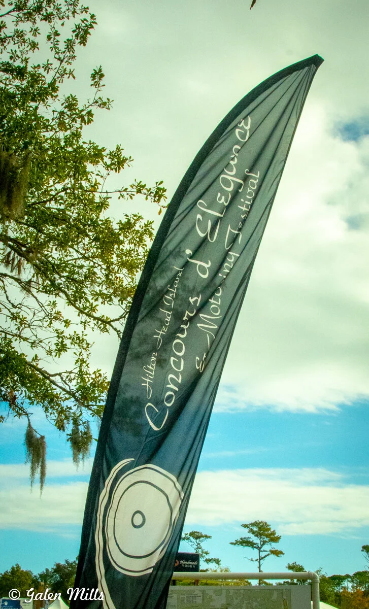 Flag reading 'Concours d'Elegance & Motoring Festival Hilton Head Island' against a backdrop of sky and trees.