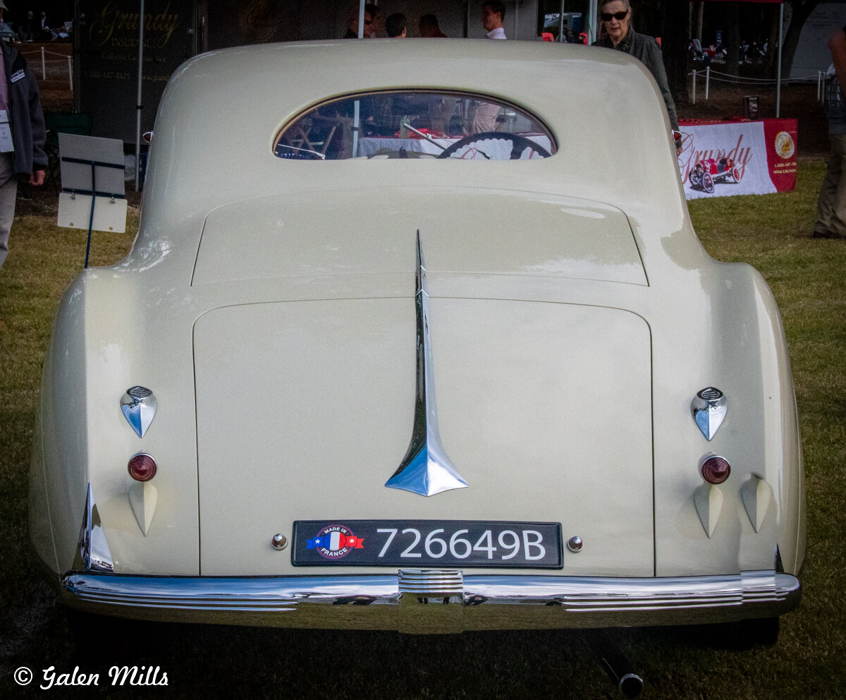 Rear view of a vintage cream-colored car with distinctive tail fins and chrome detailing, displayed at an outdoor event.