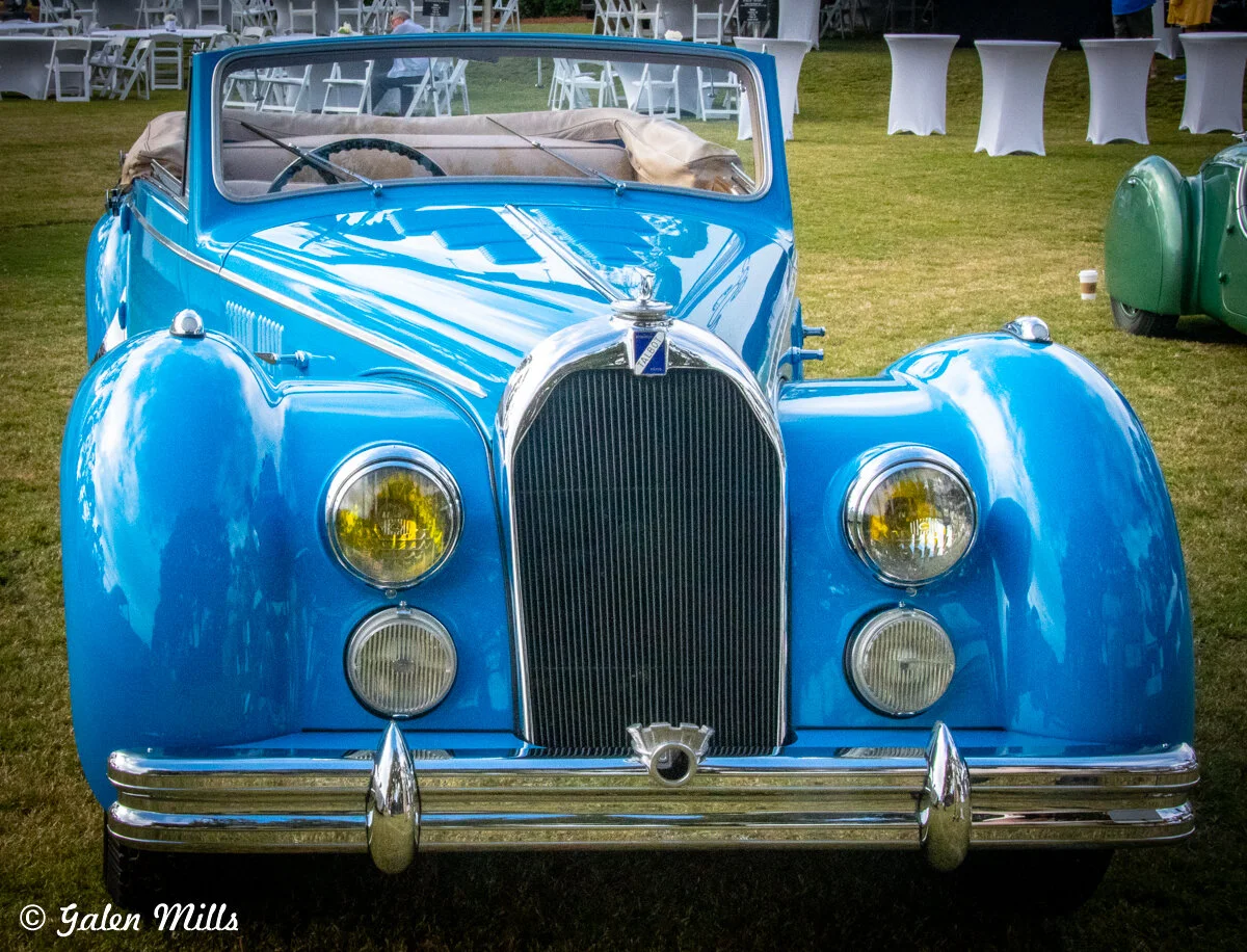 Blue vintage convertible car parked on grass with white seating in the background.