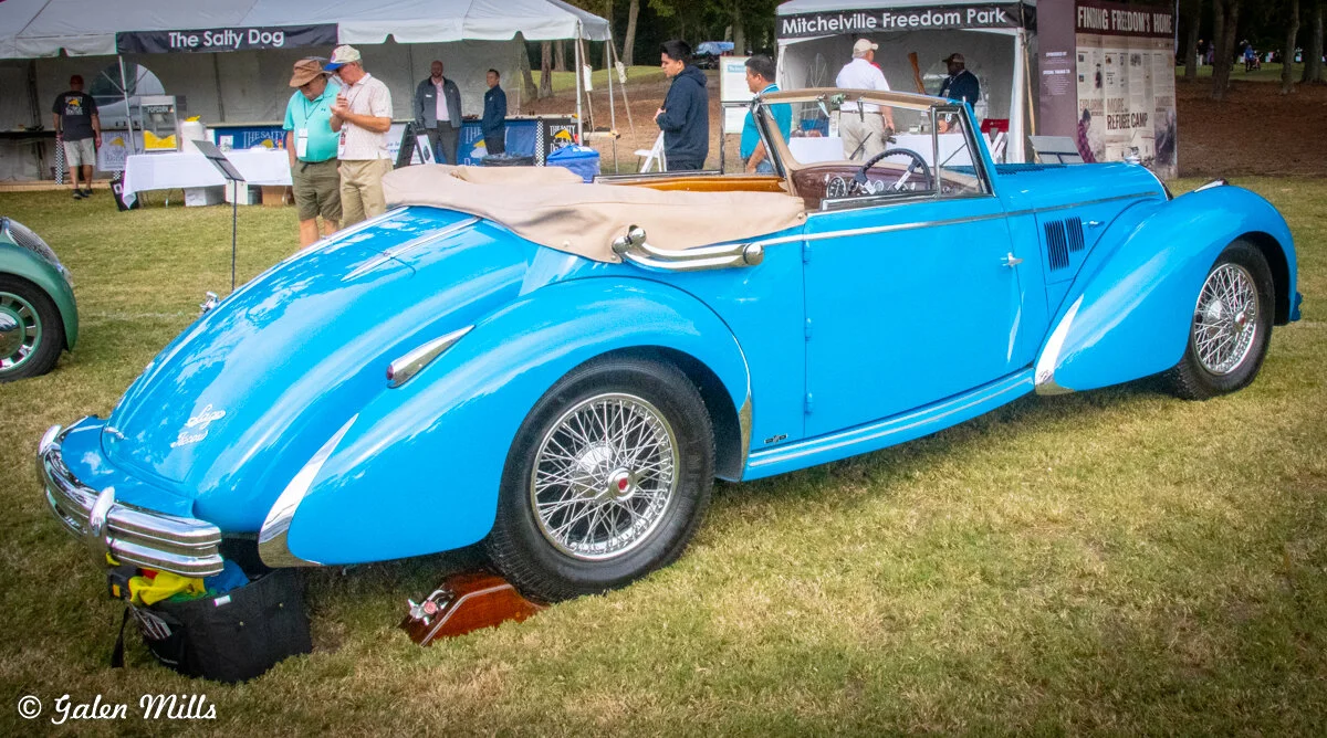 Blue vintage convertible car on display at an outdoor event, with tents and people in the background.