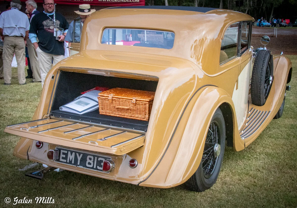 Vintage yellow car with open trunk revealing a wicker basket and a book, parked on grass with people standing nearby.