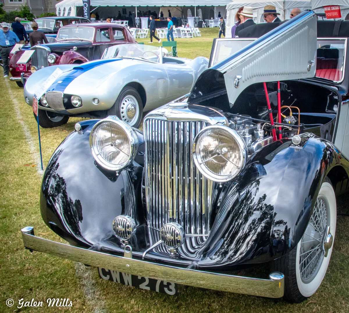 Classic car show featuring vintage cars with an open-hood black classic car in the foreground and other cars lined up in the background. Tents and people are visible in the distance.