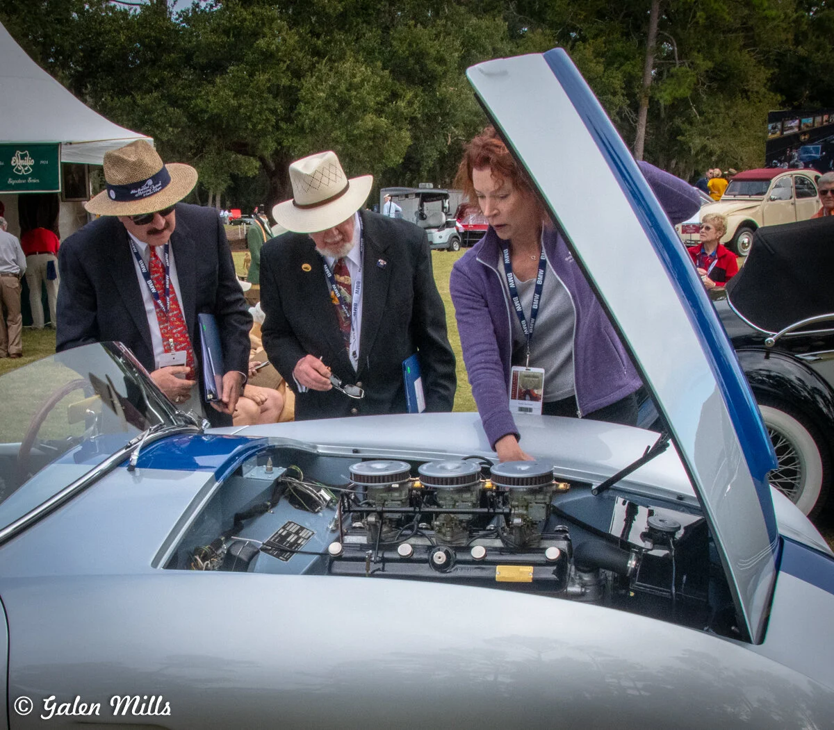 People examining a classic car engine at an outdoor car show.