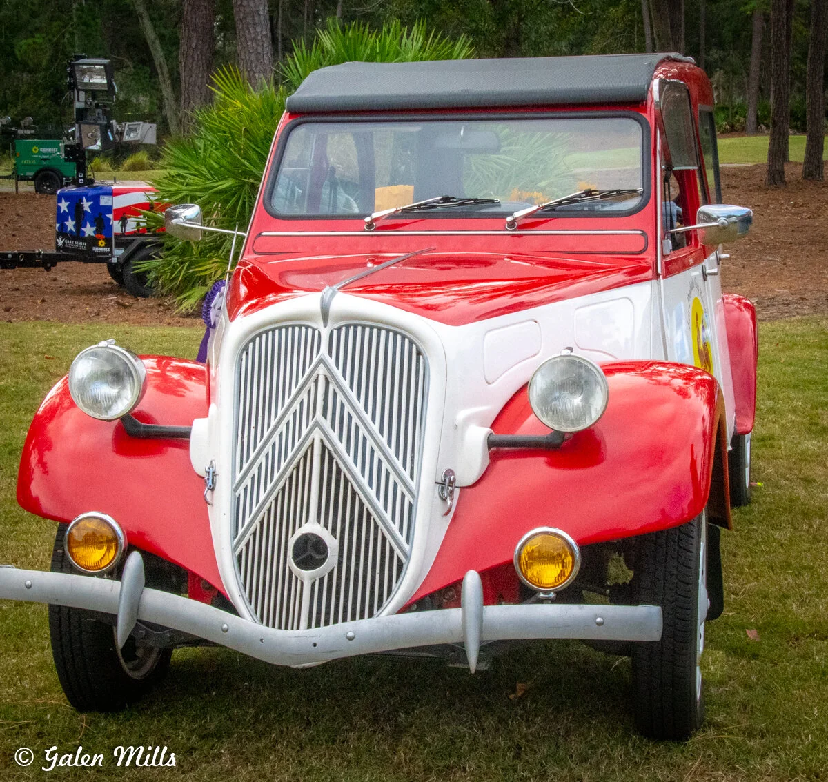 Classic red and white car with a rounded body and chrome grille parked on grass, trees in the background.