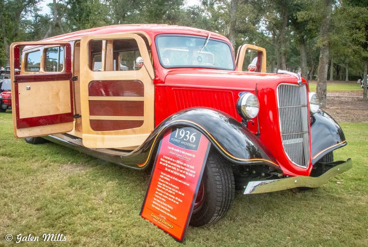 A restored 1936 Ford Woodie wagon with red and wood paneling on display, featuring its open doors and a sign detailing its history. The car is parked on grass with trees in the background.
