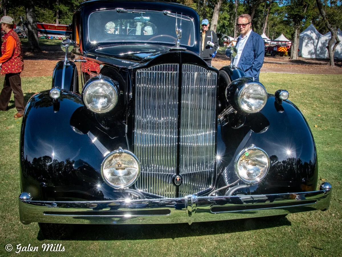 Black vintage car with shiny chrome details parked on grass at a car show, surrounded by people.