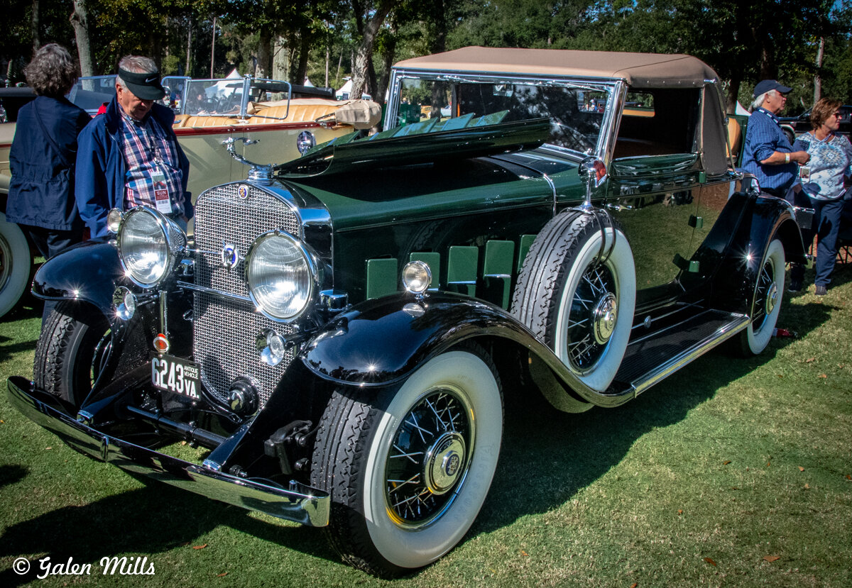 Vintage black and green classic car with a soft top, displayed at an outdoor car show with people nearby.