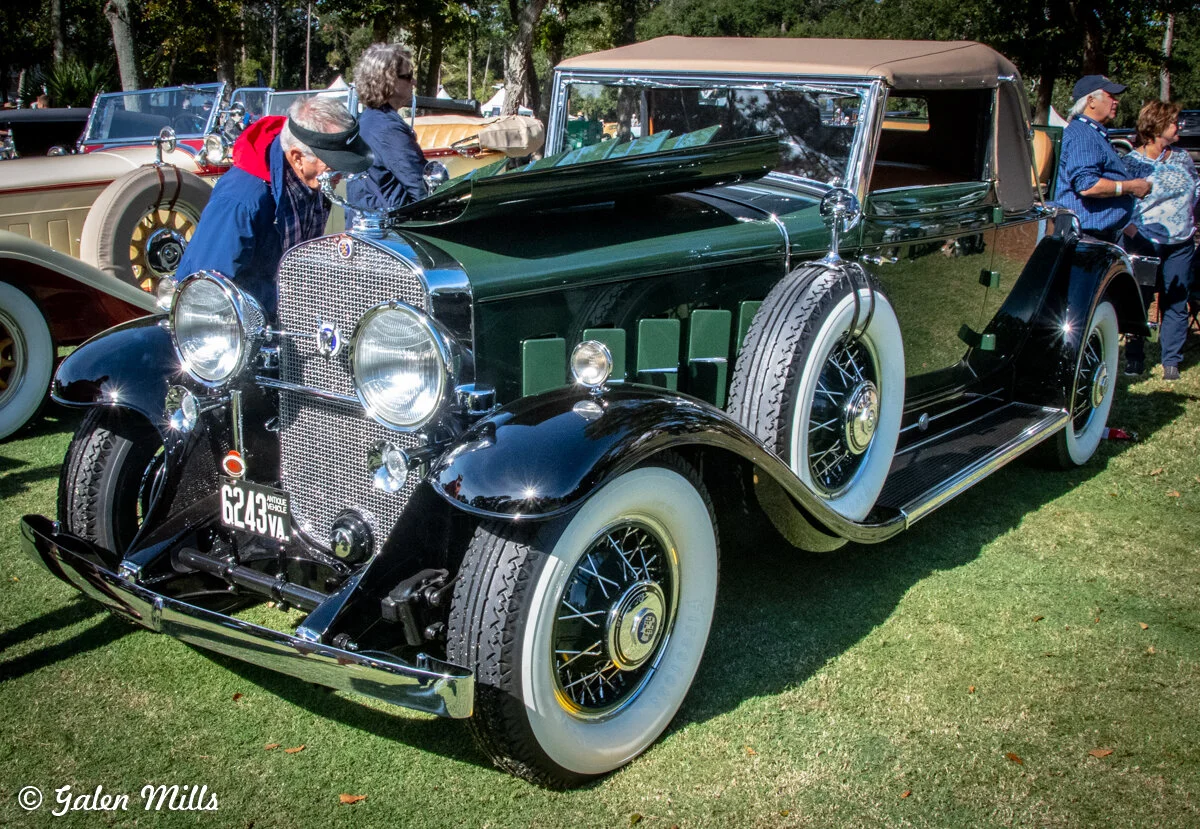 A classic vintage car with a dark green body, chrome headlights, and whitewall tires displayed outdoors during a car show.