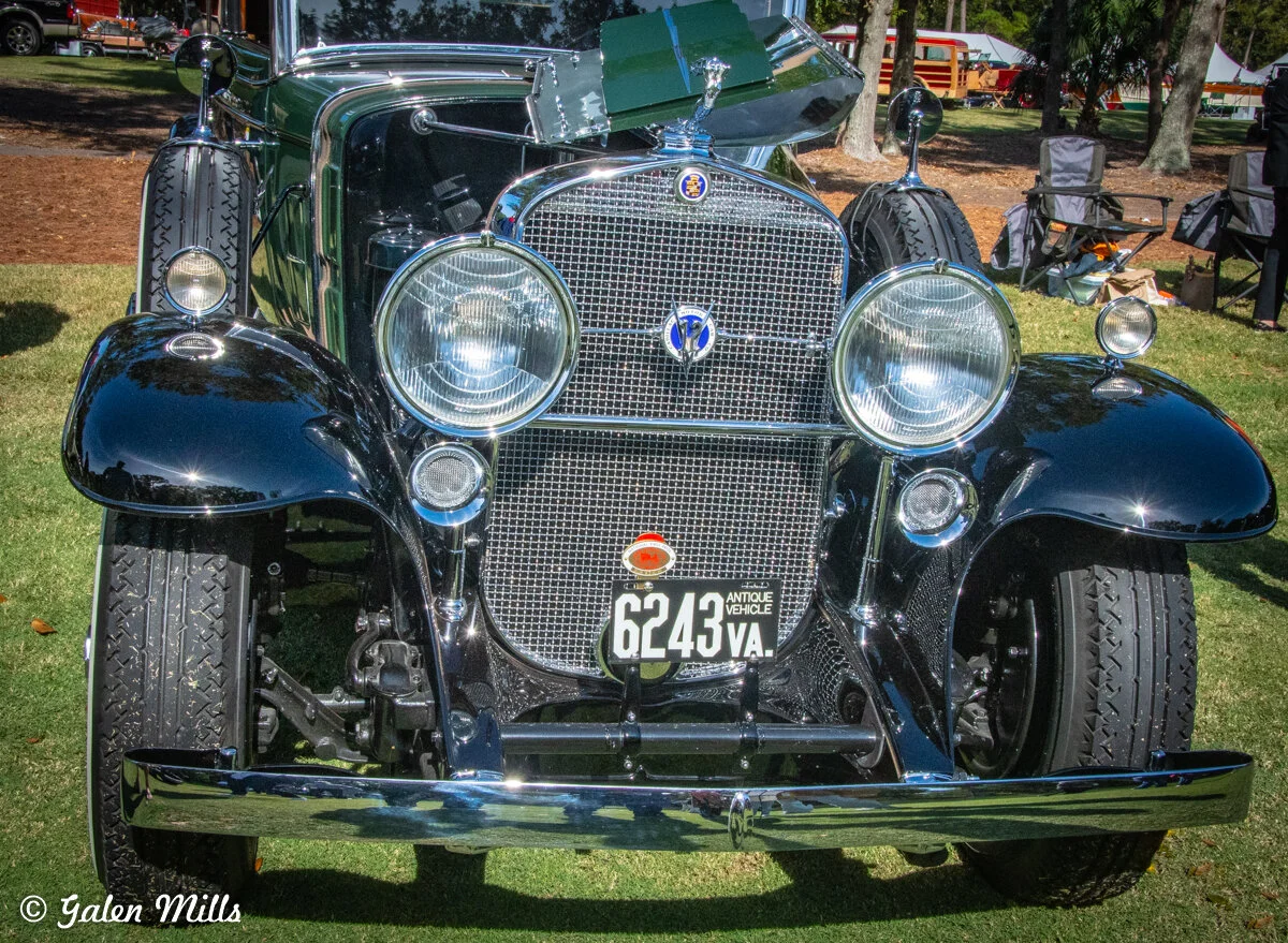 Front view of an antique car with round headlights and a classic grille at an outdoor car show.