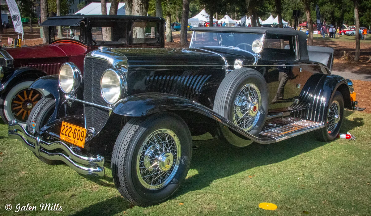 Vintage black classic car with chrome details on display outdoors; features shiny wire wheels, dual spare tires, and a 1928 Virginia license plate.