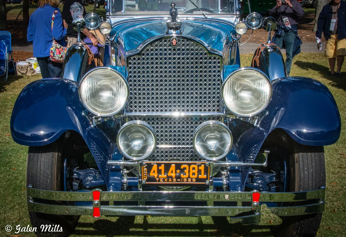 Front view of a vintage blue car with four round headlights, chrome grille, and a 1929 Texas license plate. Individuals in the background are observing the vehicle at an outdoor event.