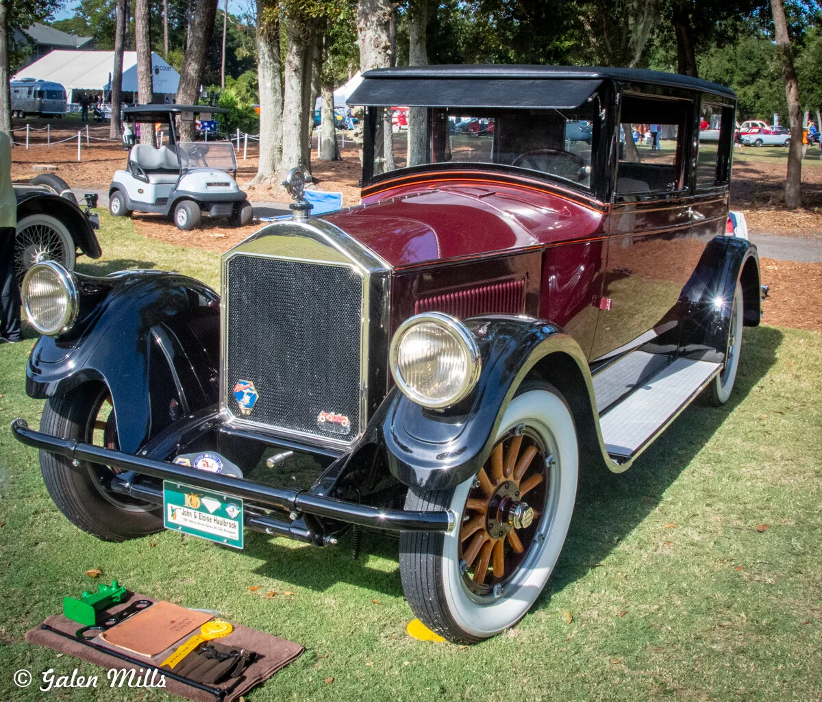 Vintage car with maroon body and black fenders, whitewall tires, displayed outdoors on grass. Nearby, a golf cart and tents are visible in the background.
