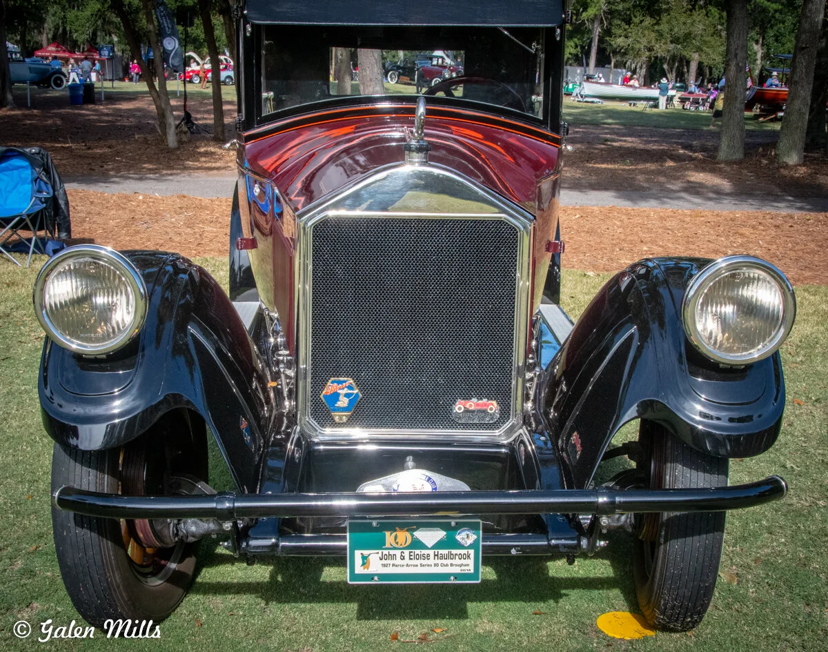 Front view of a vintage black classic car with a chrome grille, round headlights, and a nameplate. The car is on display outdoors with other vehicles and people in the background.