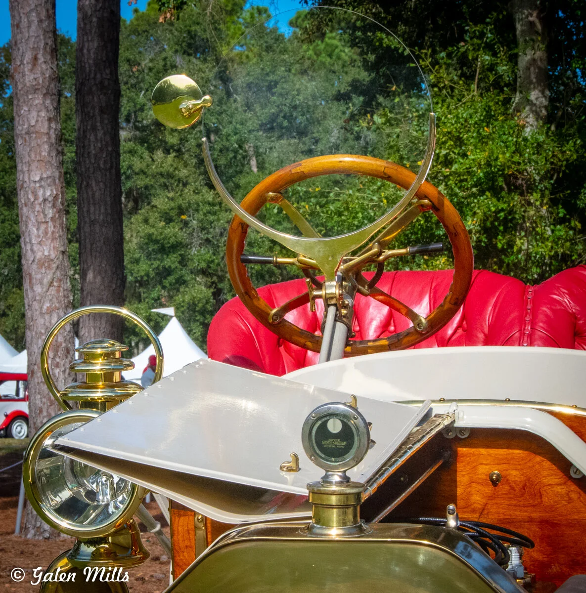 Close-up of a vintage car dashboard with a wooden steering wheel, brass details, and red leather upholstery, set against a forest background.